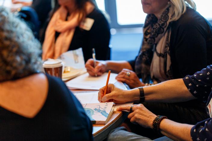 "A group in the middle of a discussion. They are sat at a table covered by paper and pens."